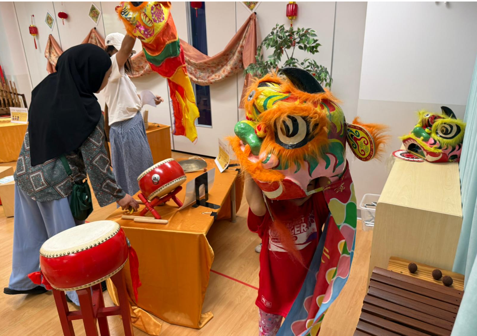 Person wearing a colorful lion dance head indoors near drums, while two others handle a dragon costume.